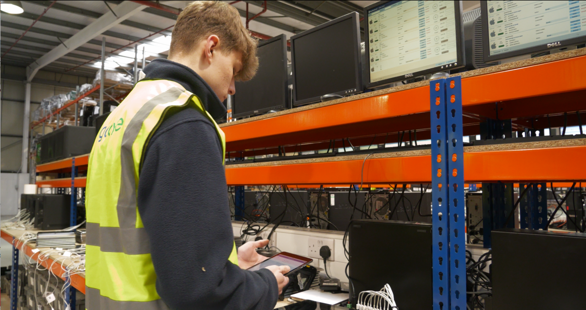 Technician in a high-visibility vest uses a tablet among server racks in a warehouse data area.