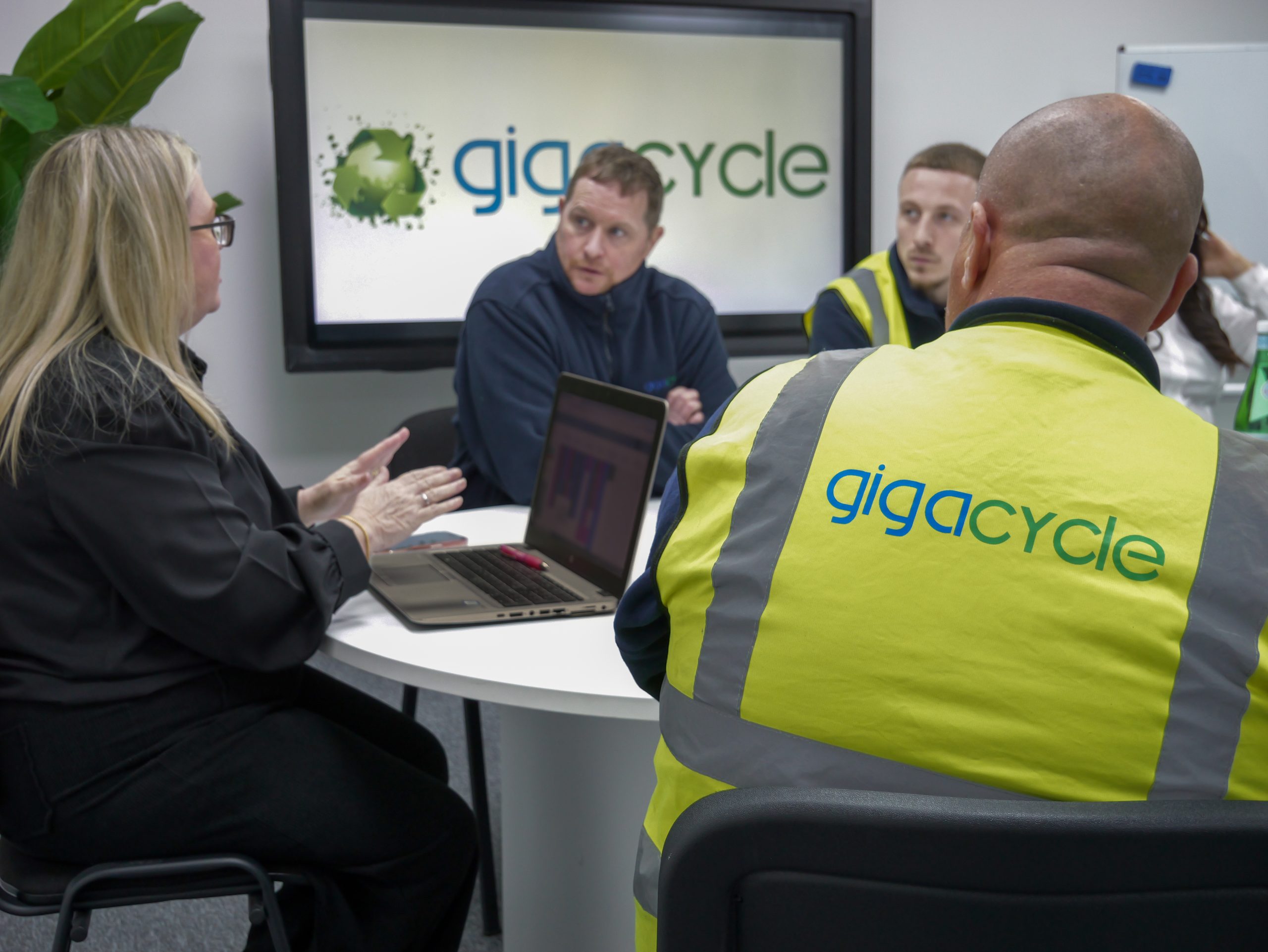 Group of professionals around a table in a discussion, one person in a high‑visibility yellow vest with 'gigacycle' logo visible in the background.