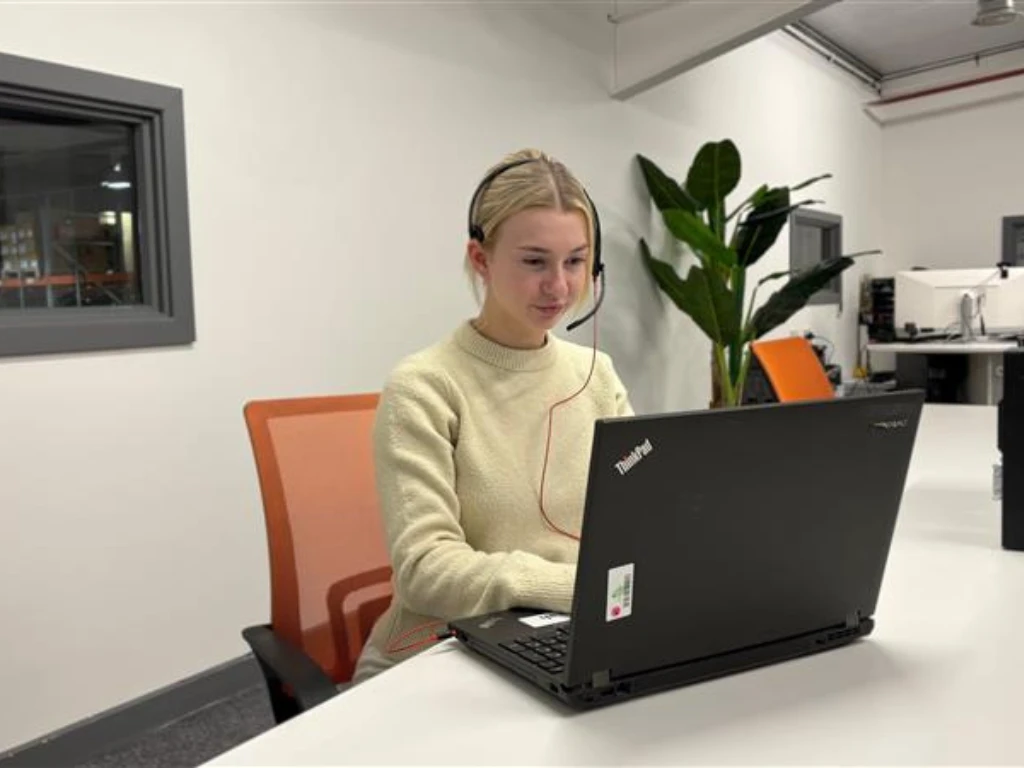 Woman with a headset working on a laptop at a white desk in an office, orange chair behind her and a tall plant in the background.