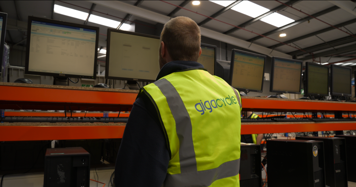 Worker in a bright safety vest seen from behind, standing in a warehouse/server room with multiple computer monitors in the background (logo reads 'gigacycle').