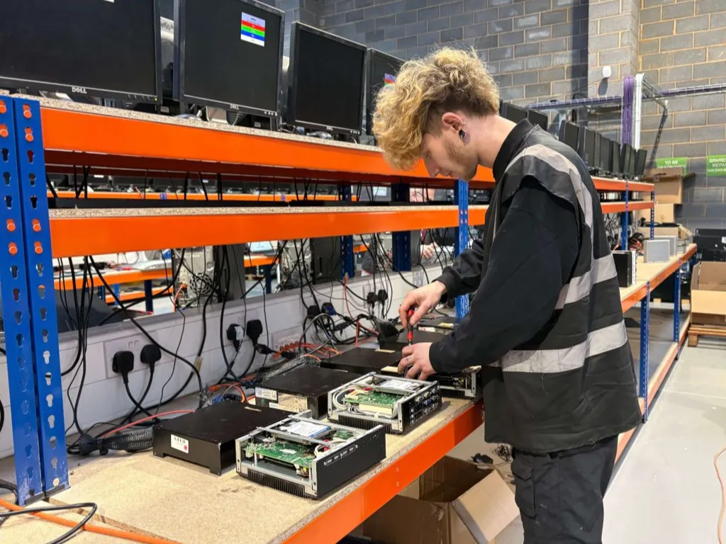 office_relocation - Gigacycle Technician in a high-visibility vest working on open server or computer hardware on a workshop bench in a warehouse setting.