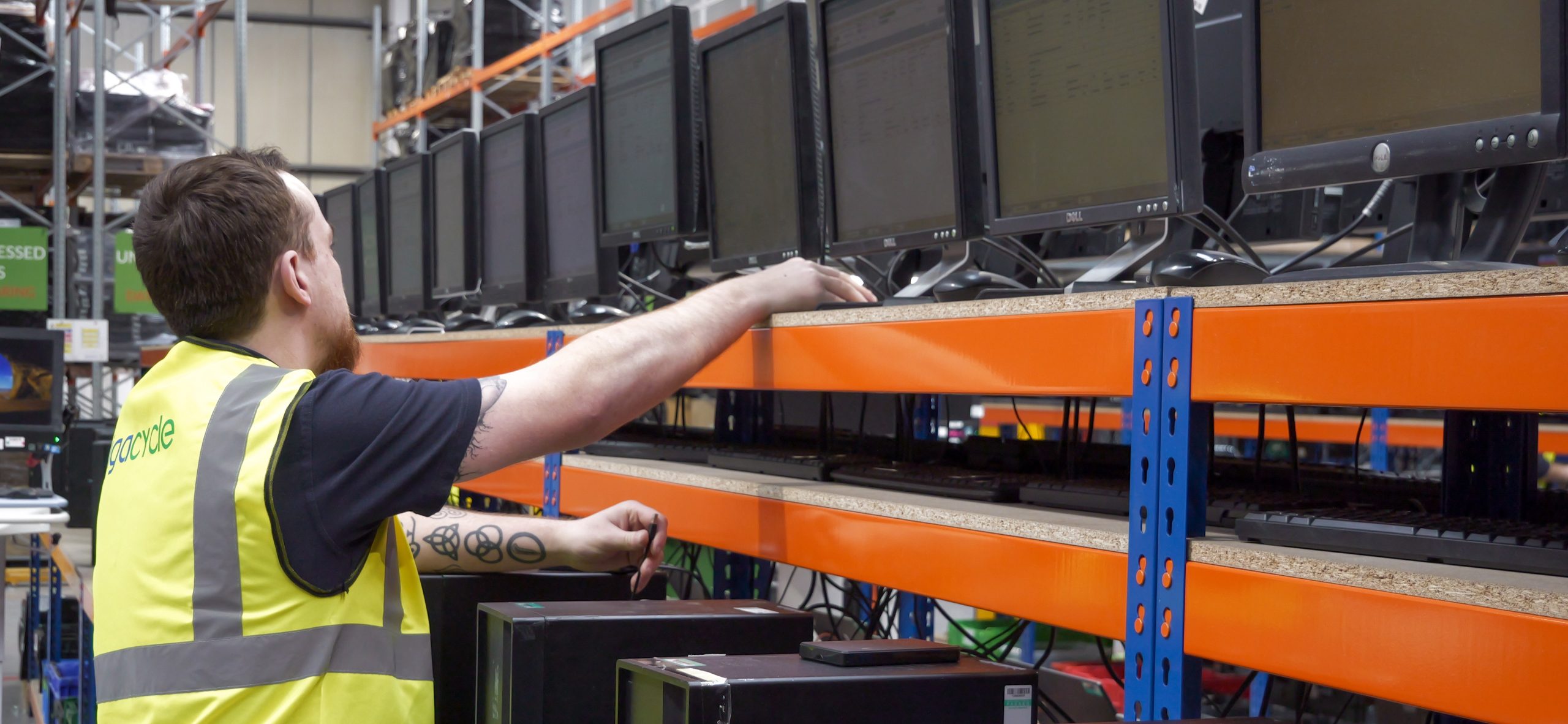 Warehouse worker in a high-visibility vest operating multiple computer monitors on orange shelving in a warehouse environment.