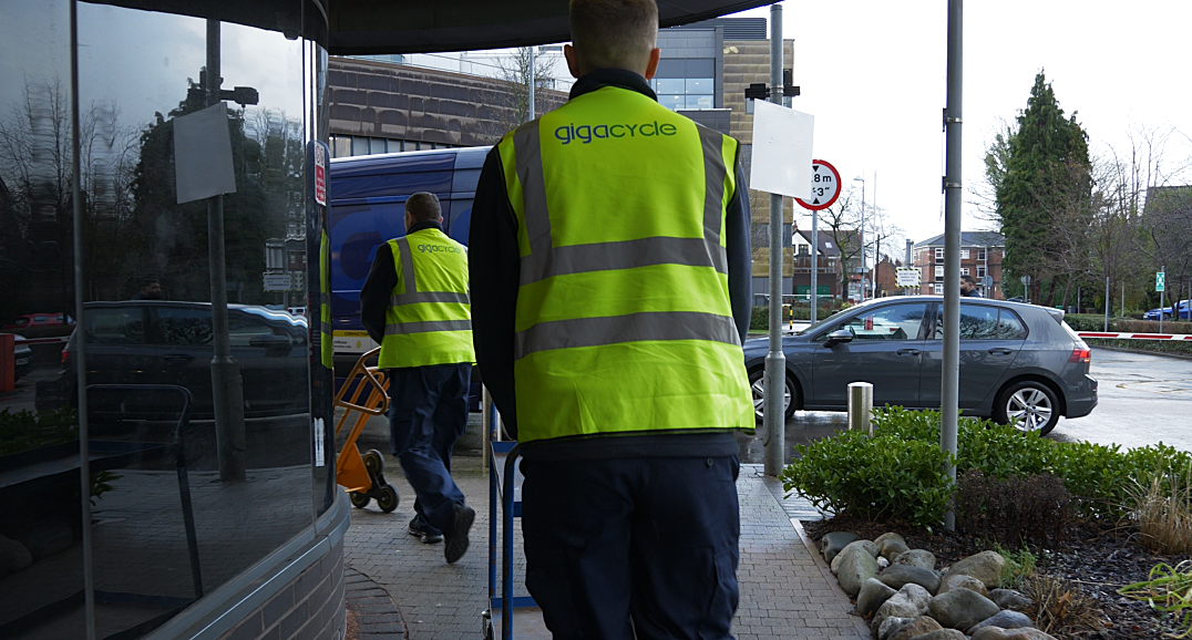 Two workers in high‑visibility vests labeled 'gigacycle' walk toward a blue truck with a hand truck nearby outside a building.