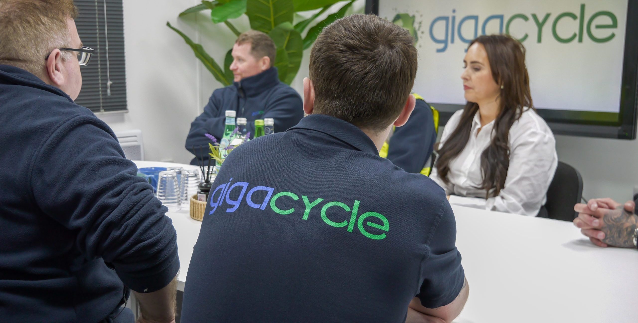 People seated around a white conference table in an office; a woman in a white blouse speaks while others listen, with a large 'gigcycle' logo on the wall.