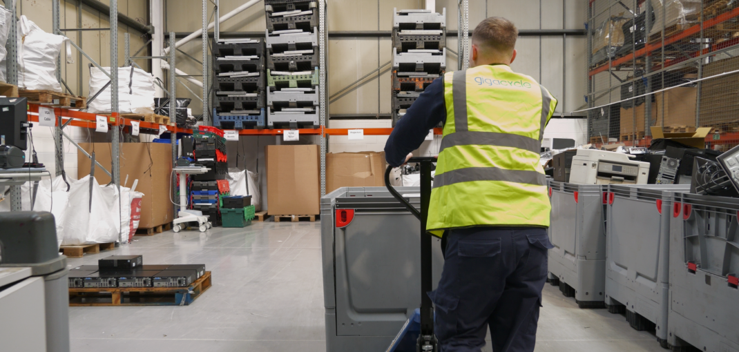 Warehouse worker in a fluorescent safety vest pushes a pallet jack down an aisle lined with shelves and pallets.