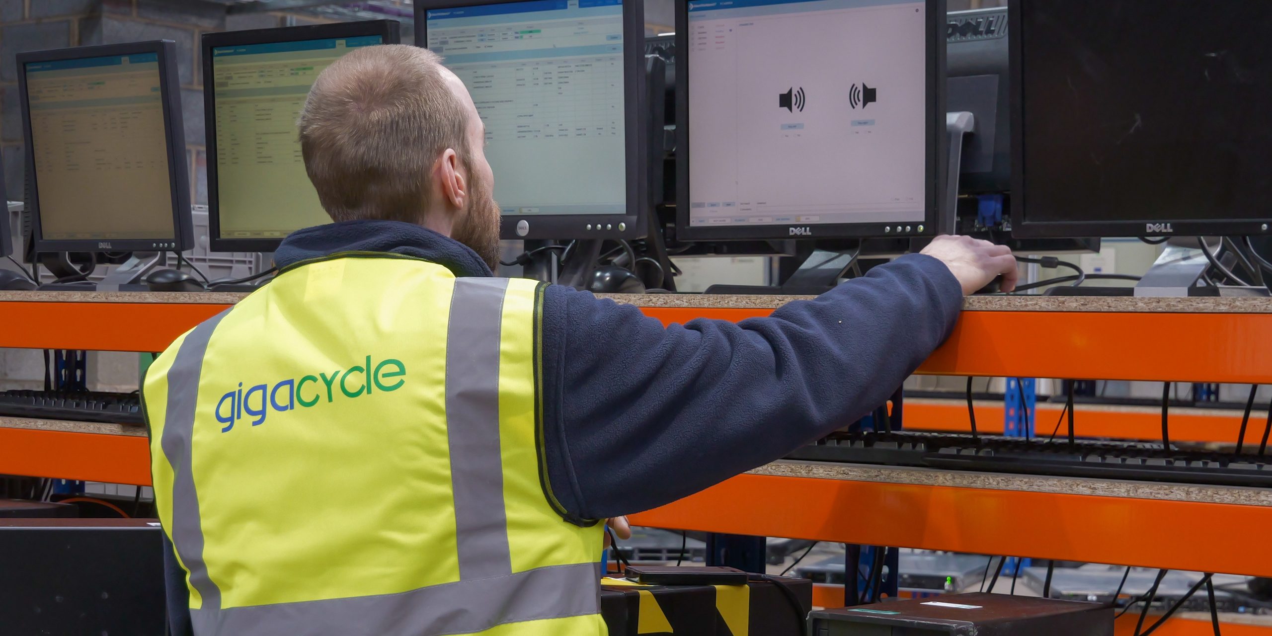 Man in a yellow reflective vest labeled 'gigacycle' works at a row of computer monitors in an industrial setup. The monitors sit on orange shelving with cables visible behind him.