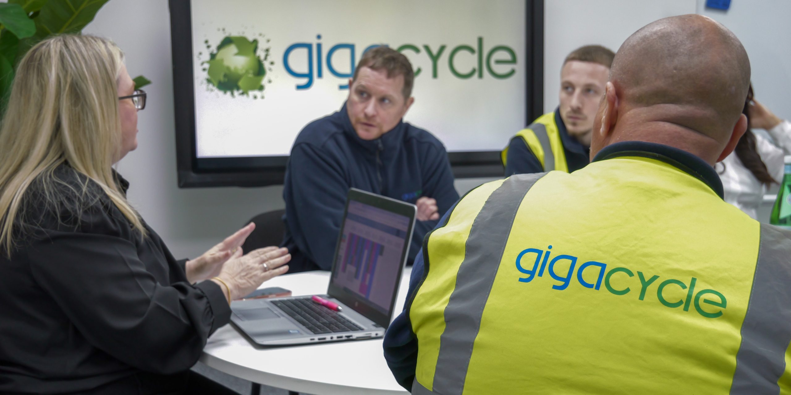 Team meeting around a table; two people in lime-vests with 'gigacycle' logo listen as a man talks, laptop with charts open nearby on a white table wall says 'gigacycle' in background