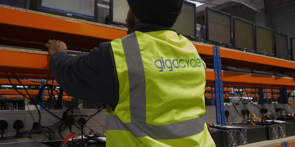 Worker in a neon safety vest with 'gigacycle' logo adjusting equipment on an orange server rack in a tech workshop. (informative)