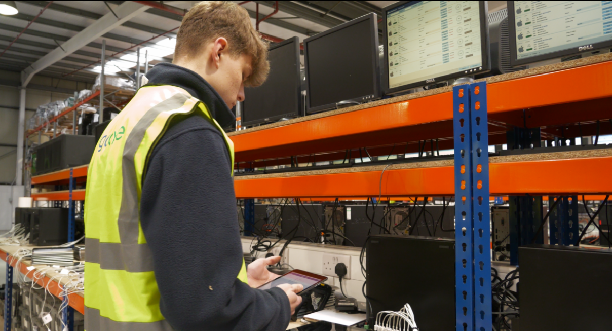 Man in a high-visibility vest uses a tablet in a data center with racks and monitors above him in orange shelving, cables around.